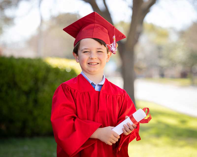 Smiling young boy in red cap and gown proudly holds diploma on sunny graduation day outdoors.