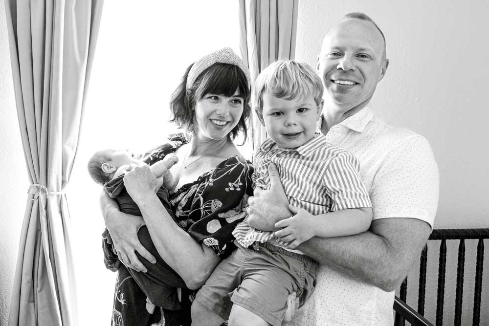 Bright lifestyle newborn family portrait with parents lovingly holding their baby and toddler by sunlit curtains.