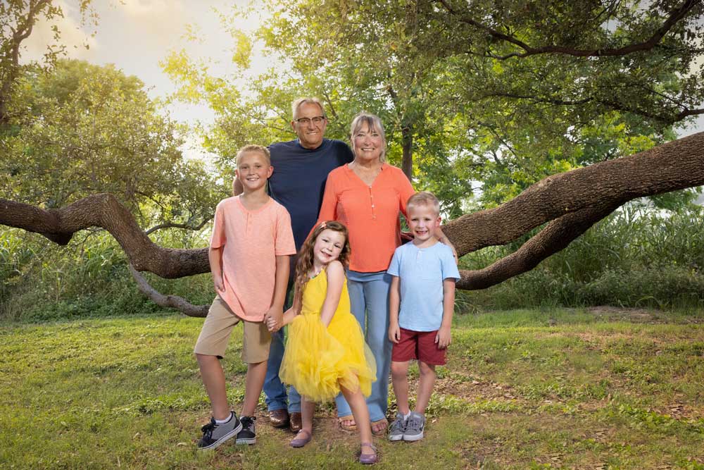 Smiling multigenerational family portraits outdoors under sprawling oak trees in a sunny, scenic park setting.