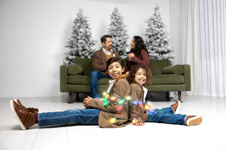 Two brothers wrapped in Christmas lights while the parents enjoy a quiet moment together on the couch. Dallas Family Holiday Portraits.